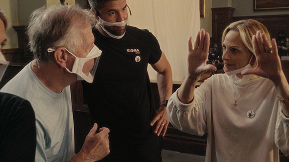 Three people stand in a room, wearing white masks pulled down below their chins.