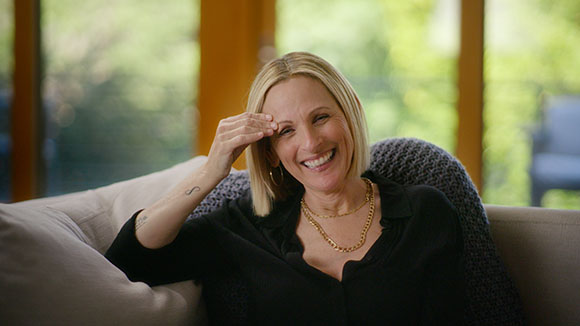A blonde woman in a black shirt laughs while sitting on a light-colored sofa.