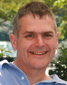 A smiling man with short greyish hair wearing a light blue button-down shirt.