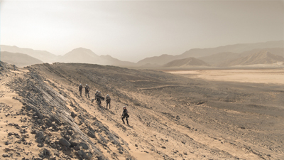 A group of people in gear hiking up a steep, rocky slope in a vast, hazy desert landscape.