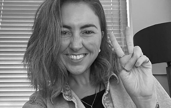 A smiling woman with shoulder-length hair holds up a peace sign in front of window blinds.