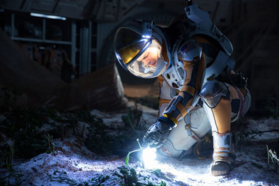 An astronaut in a white and gold suit kneels in a dark area, shining a light on small green plants.