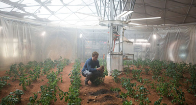 A man kneels in a large geodesic dome greenhouse, tending to small green plants growing in reddish soil.