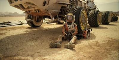 An astronaut in a white and orange suit sits on a desert landscape next to a massive multi-wheeled rover.
