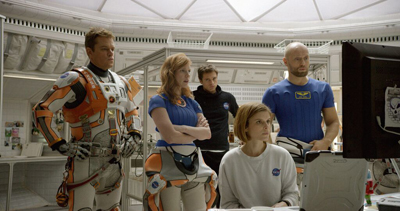 Five people in a white, high-tech space station interior looking toward a computer screen.