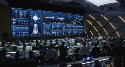 Personnel in a dark mission control room facing large blue digital data screens and monitors.