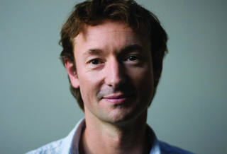 A man with wavy brown hair and a light blue shirt looking toward the camera against a plain background.