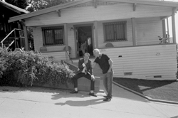 Two men pose outside a single-story house with a porch and bushes.