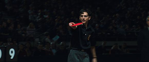 A man in a dark polo shirt holds a red paddle in a dark, crowded arena.