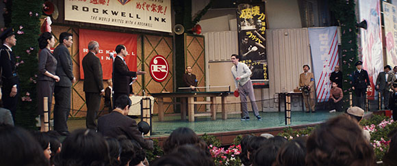 A man in a light sweater plays table tennis on a stage in front of an audience and large advertisements.