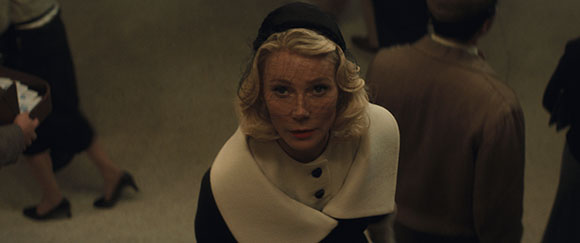 A blonde woman wearing a black hat with a veil and a white collar looks upward in a dimly lit setting.