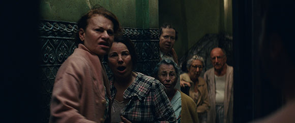 A group of elderly people in a dimly lit hallway looking toward the camera with shocked and fearful expressions.