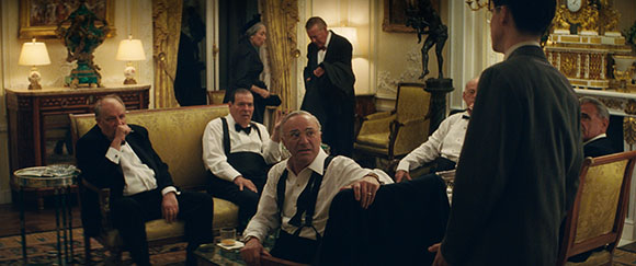 Men in tuxedos and dress shirts sit in an opulent, gold-decorated room while a waiter stands in the foreground.