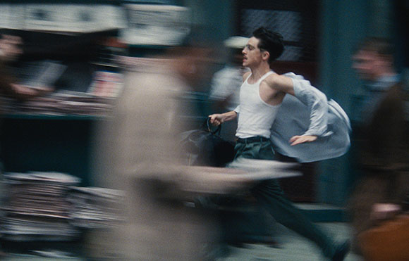 A man in a white tank top and dark pants runs through a motion-blurred urban setting.
