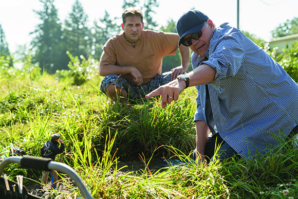 A man in a blue shirt points toward plants while another man watches from a crouched position.
