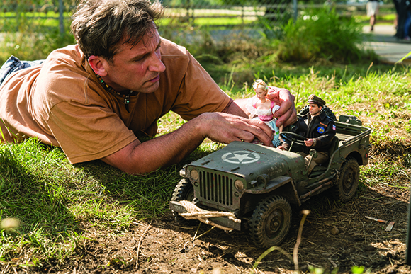 A man lying on the grass playing with a miniature military jeep and small doll figures.