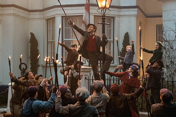 A group of people in historical clothing hold torches and long poles outside a large white building at dusk.