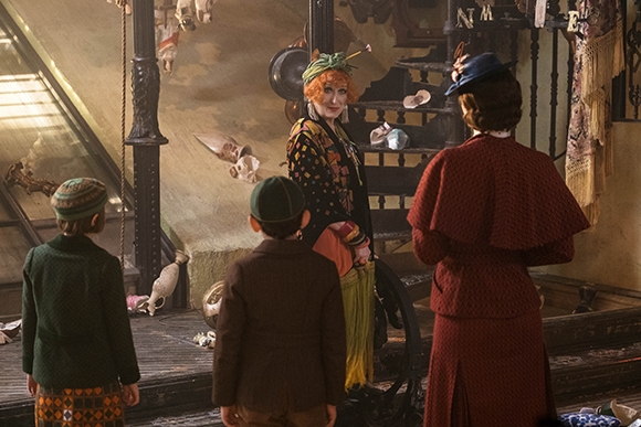 A woman in a patterned outfit and green hat stands in a room filled with curiosities facing three other people.