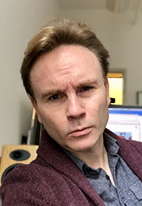 A man with light brown hair wearing a maroon cardigan over a grey shirt.