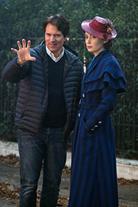 A man in a puffer jacket gestures with an open hand next to a woman in a blue dress and pink hat.
