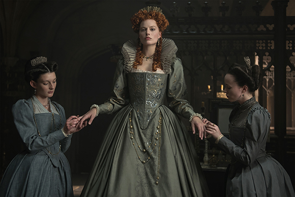 Three women in elaborate historical gowns stand in a dark, ornate room, holding hands.