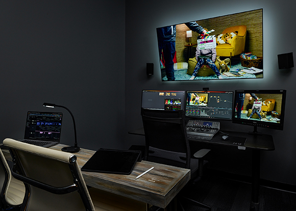 A dark editing studio featuring a large wall screen, three computer monitors, a laptop, and a digital drawing tablet.