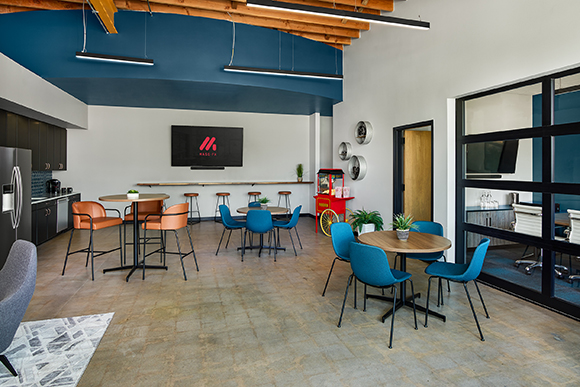 A modern breakroom with blue walls, wooden ceiling beams, round dining tables, and blue chairs.