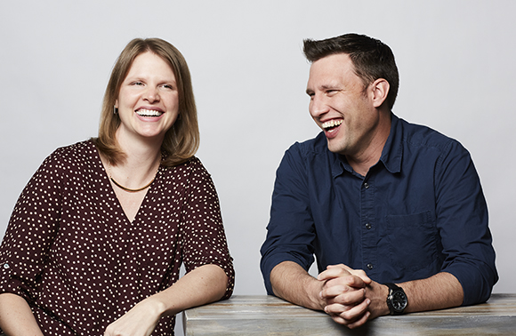 A woman in a polka dot blouse and a man in a blue button-down shirt laugh side by side against a white background.