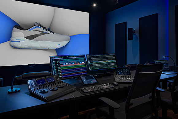 A dark video editing studio featuring multiple monitors, a control surface, and a large screen showing a shoe.