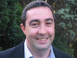 A smiling man with short dark hair wearing a dark blazer and light blue striped shirt.