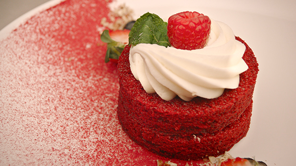A round red velvet cake topped with a swirl of white cream and a single raspberry on a white plate.