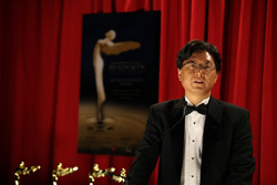 A man in a black tuxedo and bowtie speaks before a red curtain and a movie poster.