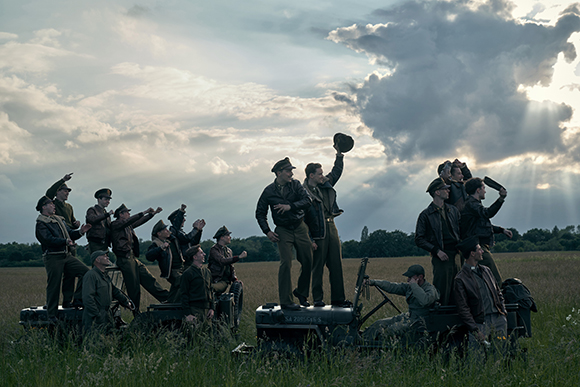 Soldiers in vintage uniforms cheering and raising arms in a grassy field under a dramatic cloudy sky.