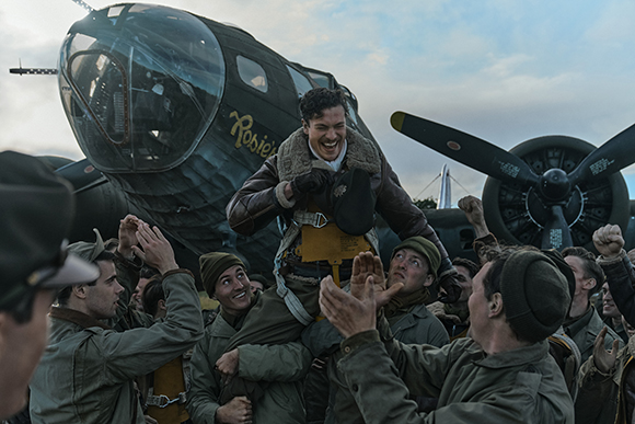 A man in a flight jacket is hoisted onto the shoulders of a cheering group of men in front of a bomber plane.