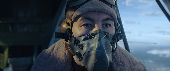 A pilot in a flight helmet and oxygen mask looking forward inside a cockpit against a blue sky.