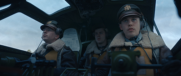 Three men in flight gear sitting inside the cockpit of an aircraft under a cloudy sky.