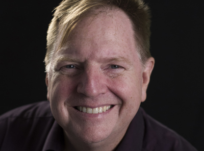 A smiling man with light hair and blue eyes looking toward the camera against a black background.