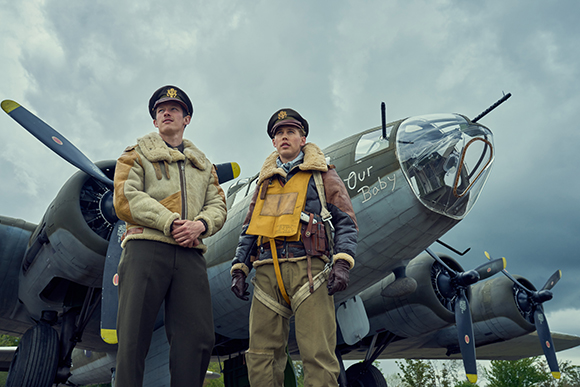Two men in WWII-era flight uniforms standing before a large multi-engine bomber under a cloudy sky.
