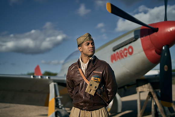 A man in a brown leather jacket and flight cap stands before a silver propeller aircraft under a blue sky.