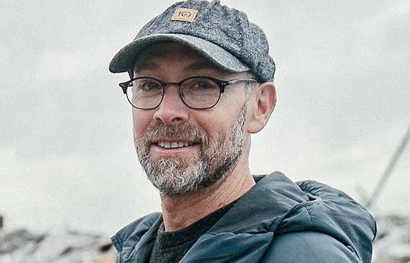 A man with a grey beard, glasses, and a grey cap smiling against a light background.
