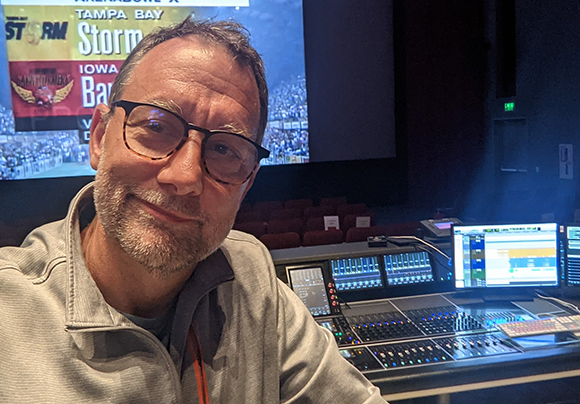 A man with glasses and a beard smiles in a dark room with a large screen and audio equipment.
