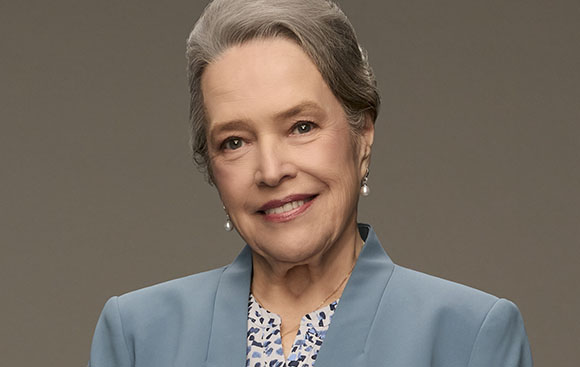 An older woman with grey hair smiling, wearing a light blue blazer and pearl earrings against a grey background.