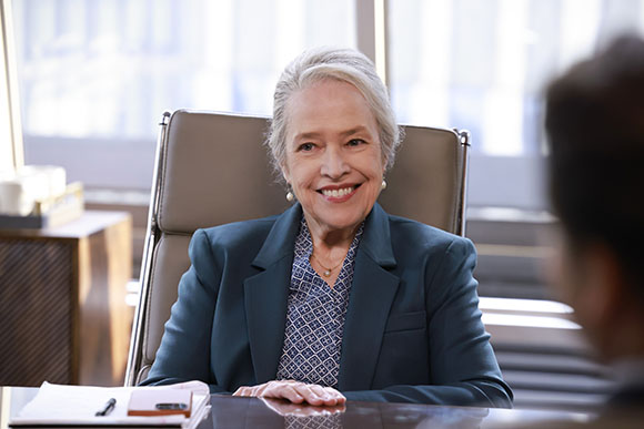 Smiling older woman with grey hair wearing a teal blazer and patterned blouse, sitting at a desk in an office.