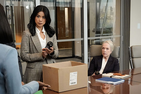 A young woman in a grey blazer holds a small black object near a cardboard box on a desk.