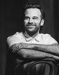 Black and white portrait of a smiling man with a beard leaning his arms on the back of a chair.