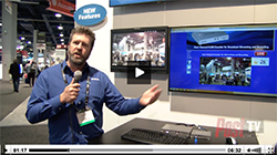 A man in a blue shirt holds a microphone and gestures toward two large digital displays at an exhibition.