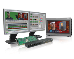 Three monitors displaying video editing software and a man's face, alongside a circuit board and hardware device.