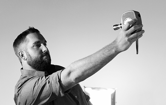 Black and white photo of a bearded man extending his arm to hold a vintage camera.