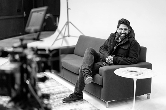 A smiling man in a jacket sits on a dark sofa in a white studio next to a small round table.