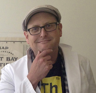 A man with glasses and a cap smiles while resting his chin on his hand.